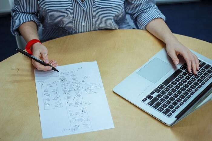 Фотография человека, сидящего за столом с компьютером и листом бумаги с рабочим процессом на нем