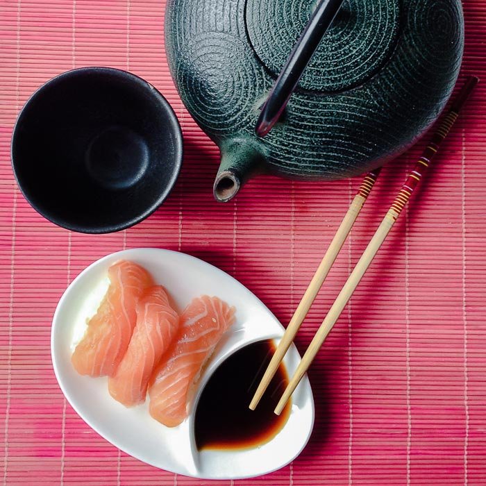 Три кусочка нигири из лосося на тарелке с палочками для еды, соевым соусом и чайником и чашкой в японском стиле рядом на столе