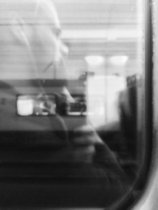 Черно-белая фотография человека в наушниках, едущего на поезде, в окне которого видно отражение другого поезда