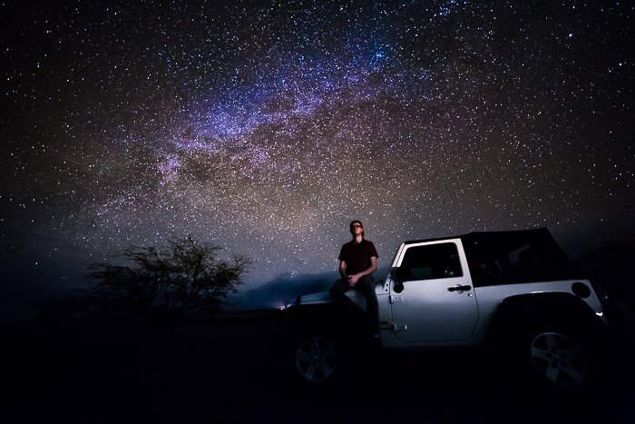 Фотограф Млечного пути сидит на капоте автомобиля под небом, усыпанным звездами