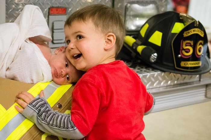 Семейный портрет, на котором маленький мальчик и новорожденный позируют с атрибутикой пожарного