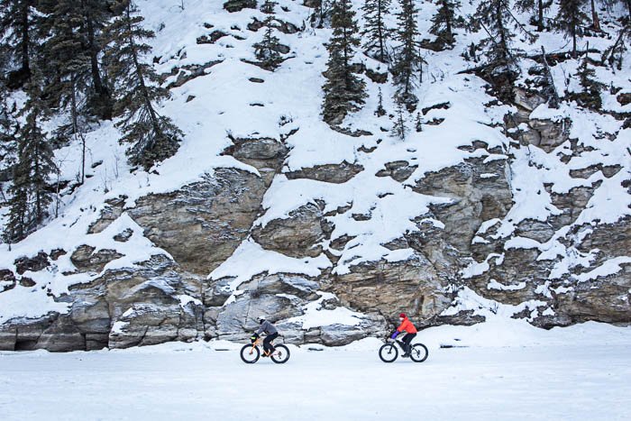 Вид издалека на двух велосипедистов, едущих по снегу. На заднем плане - горный склон.