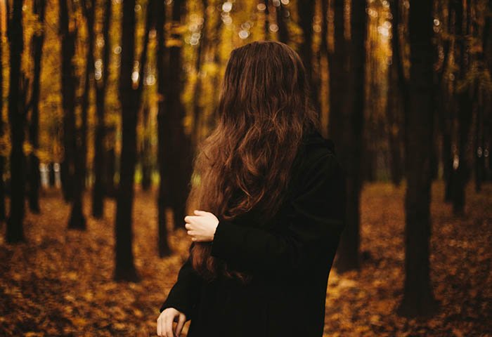 Женщина, одетая в черное, смотрит в сторону от фотографии, стоя в лесу осенью. Автопортретная фотография