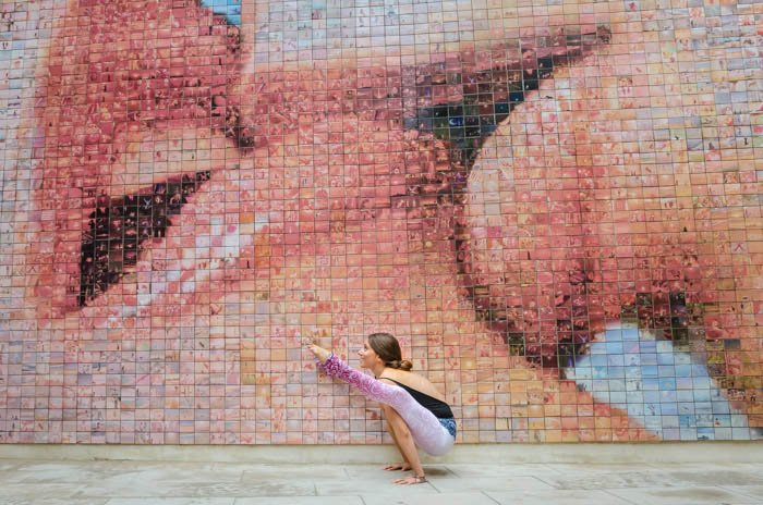 Художественная йога-фотография модели в позе светлячка перед мозаичной стеной с изображением двух целующихся людей