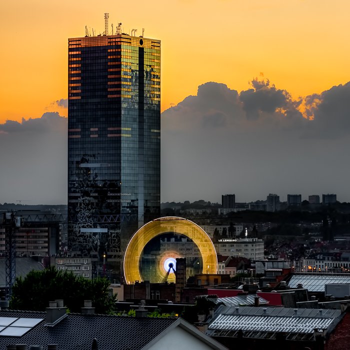 Ярко-желтое небо и облака за высокими зданиями, крыши и желтое свечение от движения колеса обозрения. Результат длинной экспозиции, снятой с 10-стопным ND фильтром.