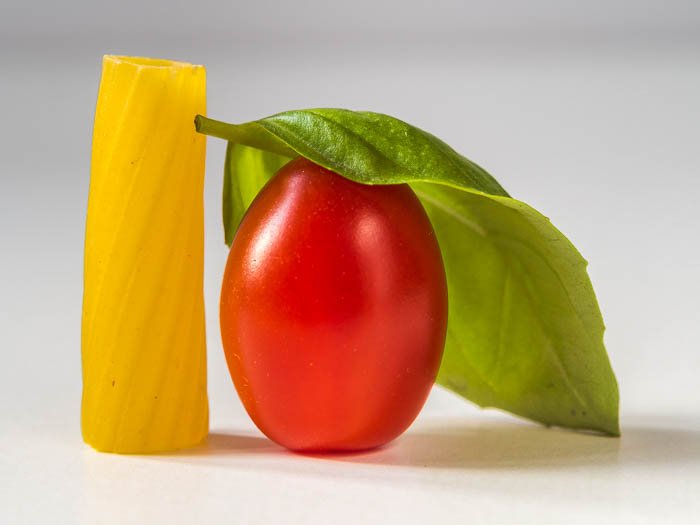 минималистское фото еды крупным планом одного куска пасты ригатони, одного сливового помидора и листа базилика 
