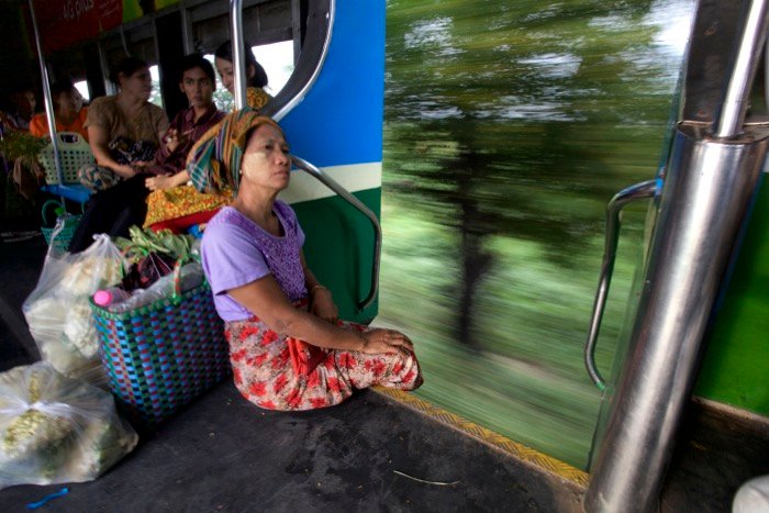 Женщина сидит на краю движущегося поезда, позади нее спины фруктов и овощей. Уличная фотография