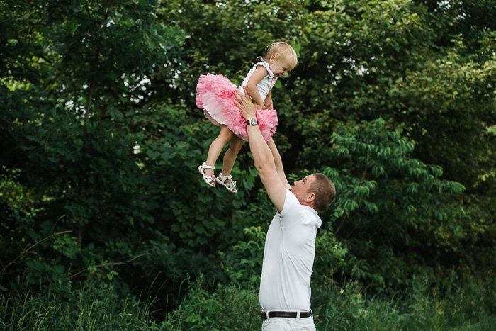 Игривый семейный портрет отца, держащего над головой маленькую девочку в розовой пачке в окружении листвы на открытом воздухе