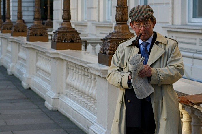 Лондонский джентльмен в кепке с козырьком и очках, курящий трубку и читающий газету на открытом городском воздухе