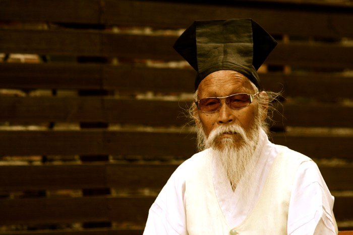 Портрет мужчины в традиционной корейской одежде и солнцезащитных очках, деревянный забор на заднем плане.