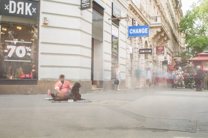 Уличная фотография с длинной выдержкой в Будапеште, размытый бездомный сидит на земле с размытыми фигурами на заднем плане.