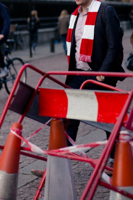 Уличная фотография мужчины в шарфе, напоминающем окружение в виде барьеров и ленты для дорожных работ. 