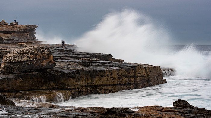 Морской пейзаж фото двух людей, рыбачащих на скалах с большими волнами позади.