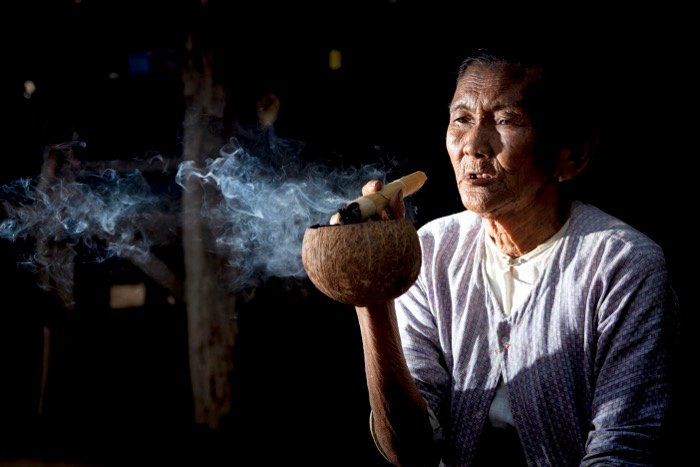 Уличная фотография пожилой женщины, курящей большую сигару и использующей скорлупу кокоса в качестве пепельницы