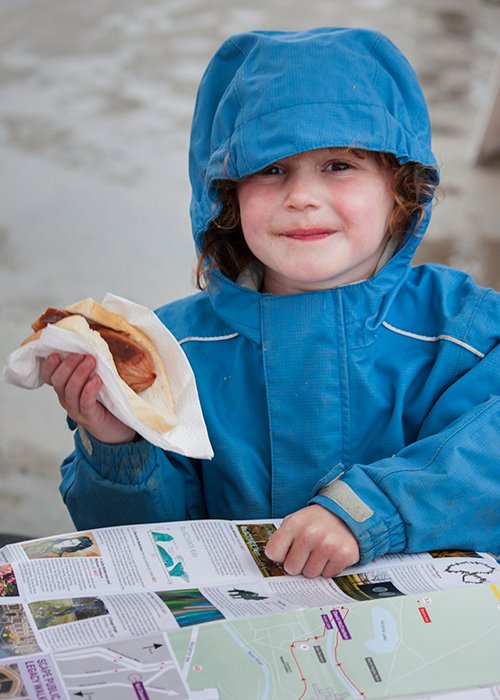 Портретная фотография маленькой девочки в синем дождевике, держащей булочку с колбасой. Советы по уличной фотографии