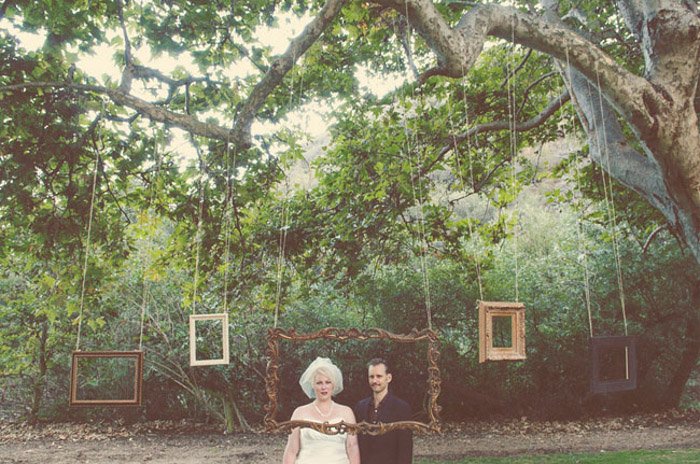 Крутая свадебная фотобудка, установленная на открытом воздухе с фоторамками разных размеров, висящими на ветке большого дерева
