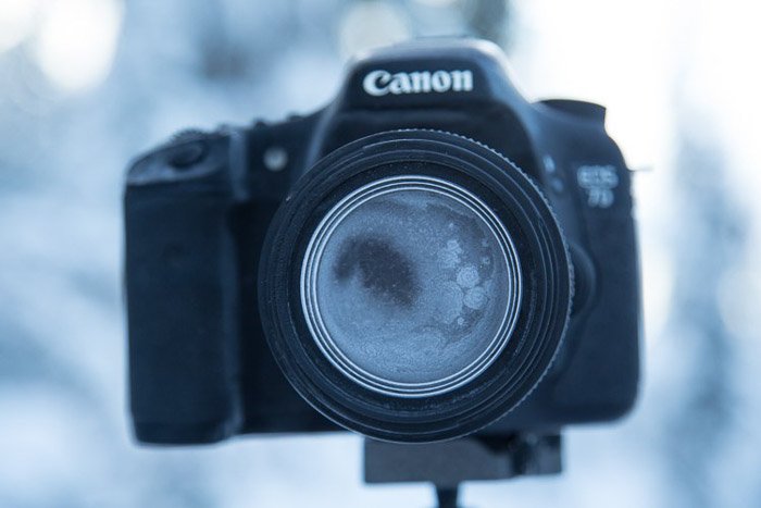 Камера Canon на штативе, объектив и корпус камеры полностью покрыты инеем, зимняя съемка.