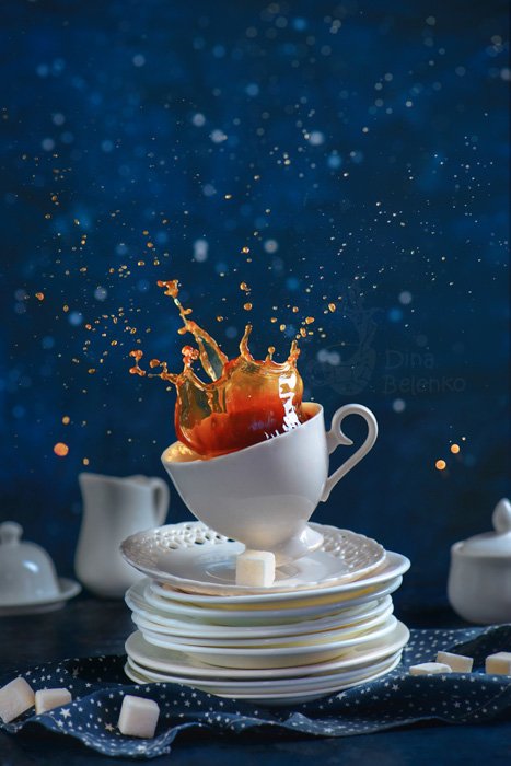 Игривая фуд-фотография кофейной чашки с брызгами, балансирующей на стопке белых блюдец на синем фоне