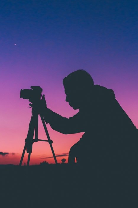 Ночной силуэт фотографа, стоящего на коленях у своей камеры на штативе, с блестящим цветным небом позади него