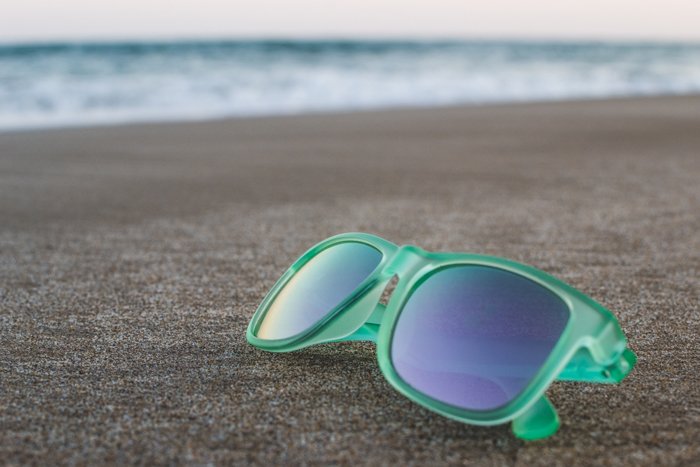 Пара солнцезащитных очков в зеленой оправе лежит на песке, морские волны размыты на заднем плане