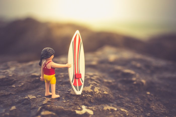 Персонаж Playmobil с доской для серфинга позирует на скалистом пляже, креативные идеи для пляжной фотографии.