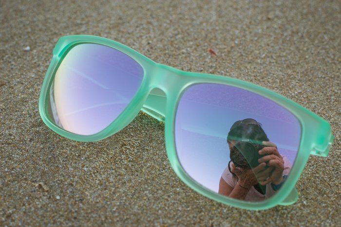 Пара солнцезащитных очков в зеленой оправе лежит на песке с отражением фотографа в одном стекле, классные идеи для пляжной фотографии. 