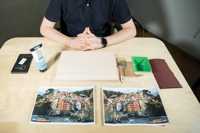 Мужчина сидит за деревянным столом, перед ним две одинаковые фотографии ярко раскрашенного прибрежного города, клей, кисть, наждачная бумага и другие материалы для переноса лазерной печати на дерево