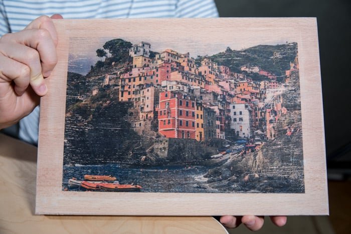Мужчина держит деревянную доску с перенесенной на нее красочной фотографией прибрежного города