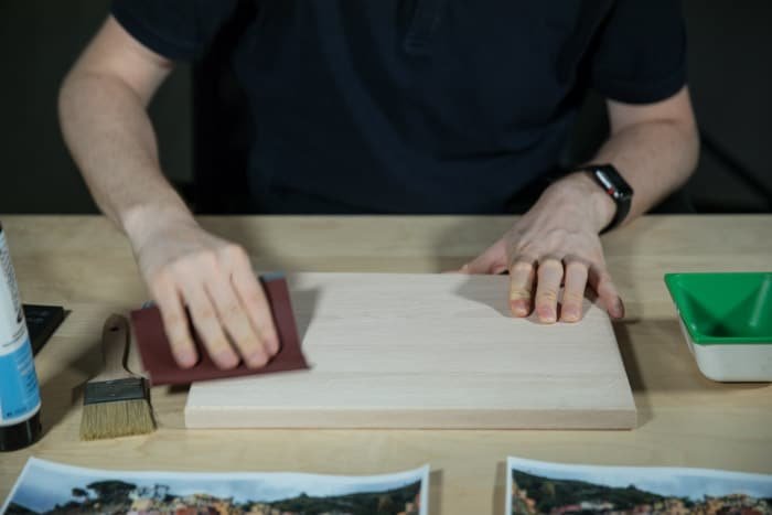 Мужчина сидит за деревянным столом, шлифуя деревянную доску, чтобы подготовить поверхность для переноса фотографии на дерево