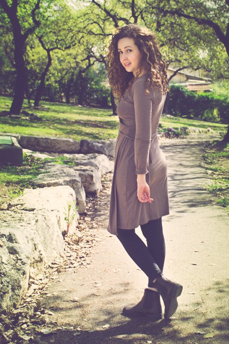 кудрявая брюнетка в коричневом платье и сапогах смотрит назад, идя по грунтовой дорожке в парке - обрезка фотографий