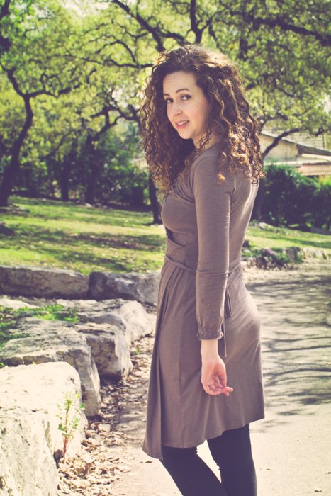 обрезанное фото кудрявой брюнетки в коричневом платье и сапогах, оглядывающейся назад, когда она идет по грунтовой дорожке в парке