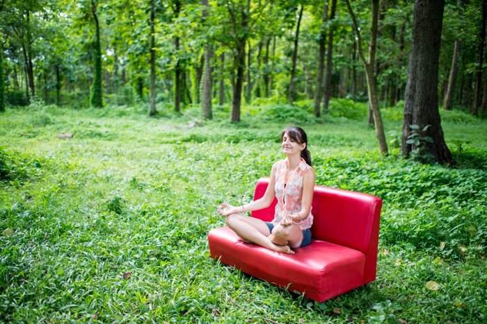 Женщина сидит на красном диване в лесу и медитирует, хороший пример редакционной портретной фотографии.