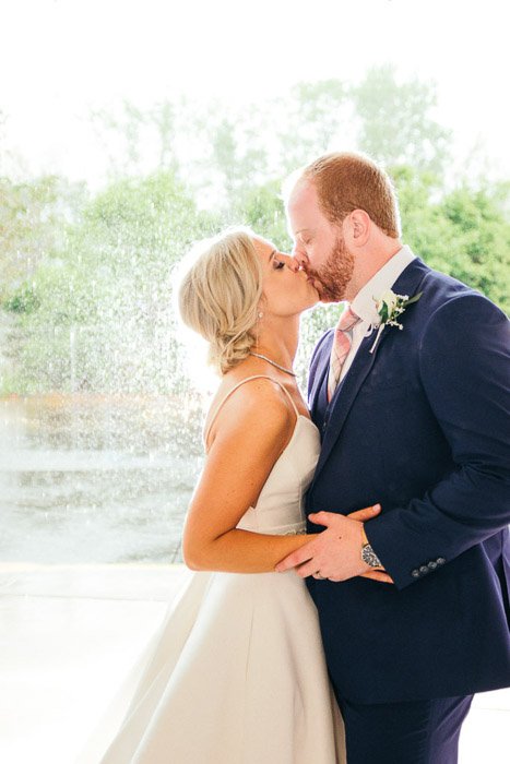 A wedding portrait of bride and groom kissing outdoors in front of a fountain