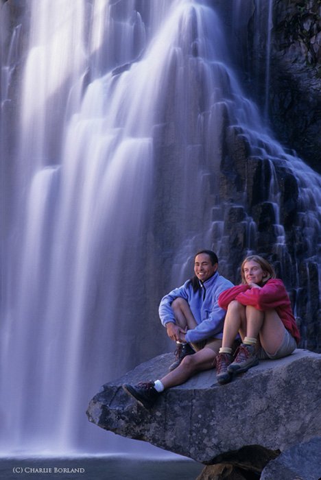 два туриста сидят на скале перед водопадом