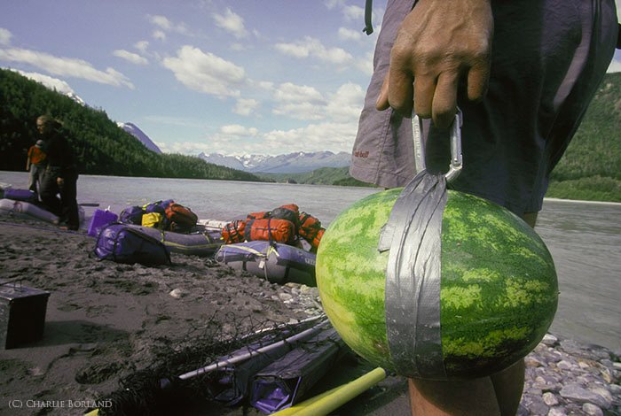 close up of the man's hand carrying a watermelon, travellers gear and the short of a lake in the background