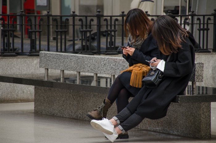 Уличная фотография двух девушек, сидящих на скамейке и пользующихся смартфонами, демонстрирующая средние и темные тона в изображениях