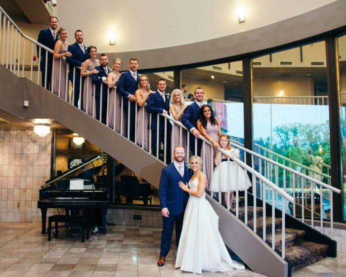Большая групповая фотография свадебной свиты, позирующей на большой изогнутой лестнице, жених и невеста внизу впереди, рядом рояль