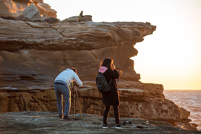 мужчина и женщина стоят у скалы, устанавливая камеру и штатив для съемки морского пейзажа