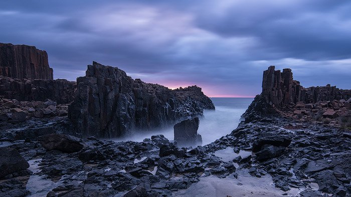 скалистый берег моря, уходящий в пенное туманное море, облачное голубое и розовое небо во время восхода солнца