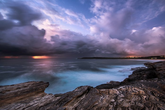 Фотография морского пейзажа на длинной выдержке в сумерках. Синее море со скалистым берегом, отражающим фиолетовое небо и облака, оранжевый закат на горизонте