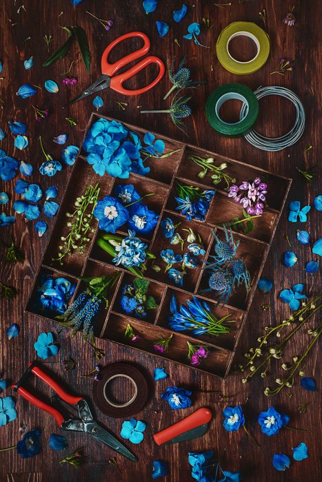 небесно-голубые цветы, розовые и фиолетовые лепестки и листья в деревянной коробке, больше лепестков и листьев на фоне деревянного стола, с ножницами, щипцами и лентой