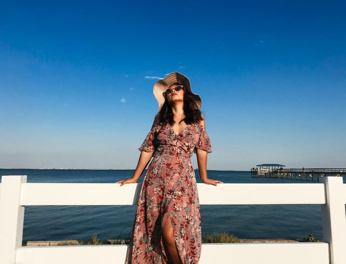 женщина в летнем макси-платье, солнцезащитных очках и большой шляпе от солнца, опирающаяся на белый забор, синее море и небо за ней