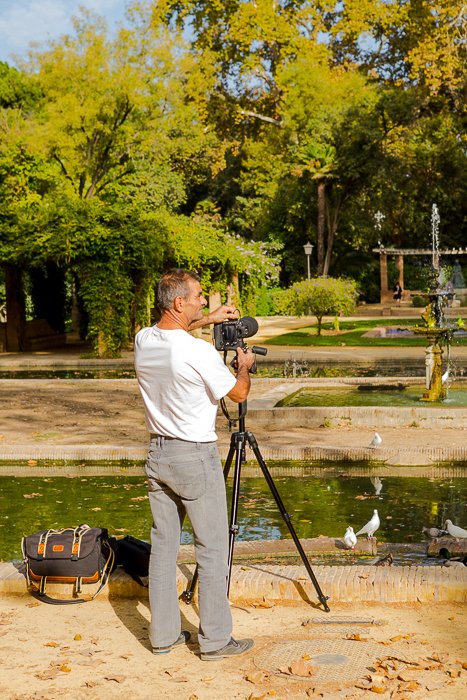 фото сзади мужчины, устанавливающего зеркальный фотоаппарат на штатив у пруда с белыми птицами, деревья вдали - советы по безопасности путешествий