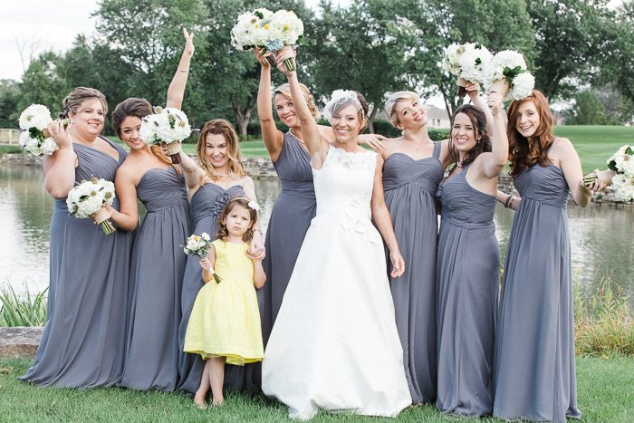 невеста с цветочницей в желтом и подружки невесты в серых платьях стоят на фоне озера, улыбаясь и держа букеты