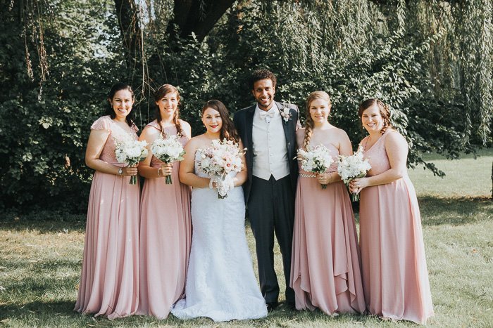 desaturated photo of bride, groom, and bridesmids in pale pink standing against the backdrop of trees.