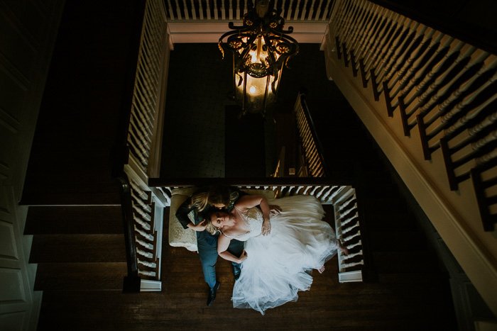 Вид с вершины лестницы на невесту, лежащую на коленях у жениха на диване, комната тускло освещена винтажной люстрой
