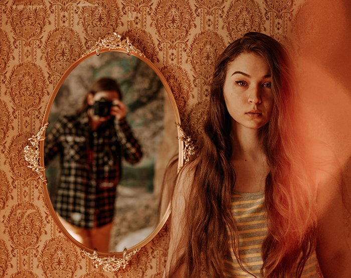 Художественный портрет в помещении женской модели, позирующей у зеркала, в котором отражается фотограф, примеры отличных фотографий профиля