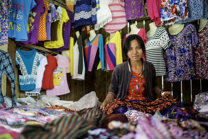 Женщина-продавец на рынке у своего прилавка, на фоне беспорядочно разбросанных цветных материалов и одежды