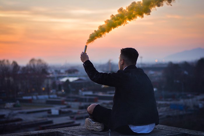 Мужчина сидит на крыше и держит желтые дымовые гранаты для фотографирования