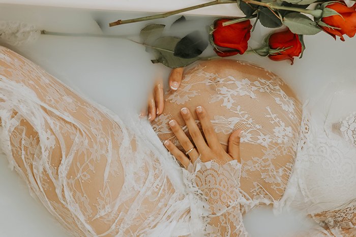 Крупный план фотосессии Milk bath беременной женщины-модели в белом кружеве, держащей свой бугорок ребенка, в окружении цветов